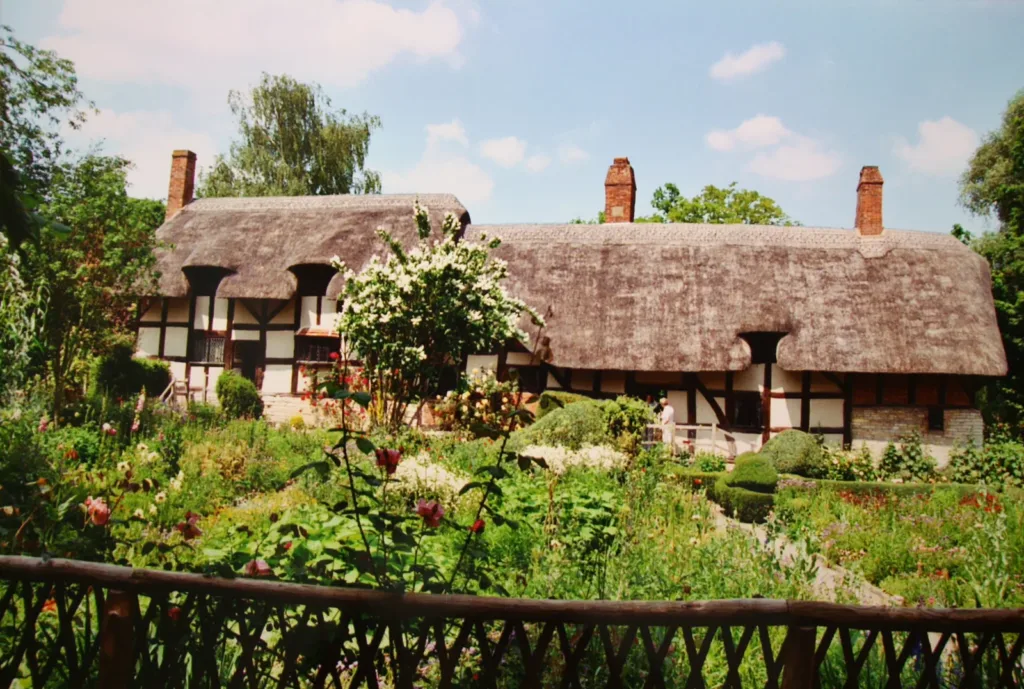 Cottage a graticcio con tetto di paglia, circondato da un giardino di fiori e bosso potato, sotto un cielo d’estate